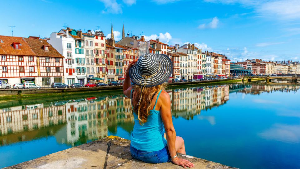 Femme assise au bord de l'eau à Bayonne, admirant les façades colorées des bâtiments qui se reflètent dans l'eau sous un ciel bleu.