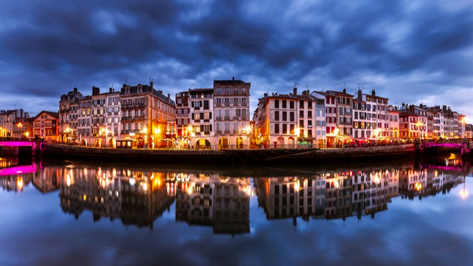 Reflets lumineux des maisons colorées de Bayonne le long de l'Adour pendant la nuit, créant une atmosphère magique et sereine.