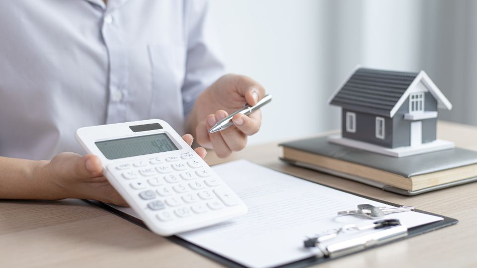 Personne utilisant une calculatrice avec un modèle réduit de maison sur une table, symbolisant le calcul des prêts immobiliers.