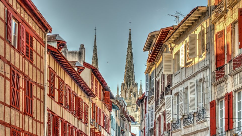 Vue d'une rue pittoresque de Bayonne avec des maisons aux volets rouges et blancs typiques de la région. En arrière-plan, on aperçoit les flèches de la cathédrale.