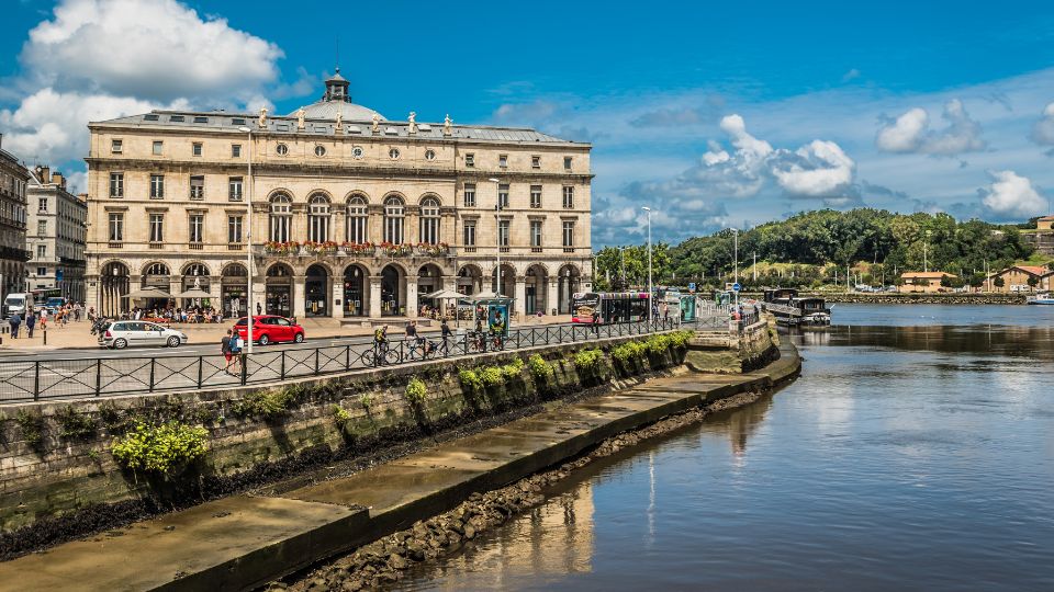 L'Hôtel de Ville de Bayonne situé le long de la Nive, avec des personnes et des voitures passant devant, sous un ciel bleu parsemé de nuages blancs.